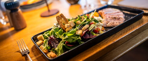 Steakgericht mit Salat und Rotwein auf schwarzem Teller Nahaufnahme eines angerichteten Gerichts mit geschnittenem, gewürztem Steak, gemischtem Salat mit Cherrytomaten, Croutons und Dressing auf einem schwarzen Teller. Im Vordergrund ein Glas Rotwein auf einem Holztisch.