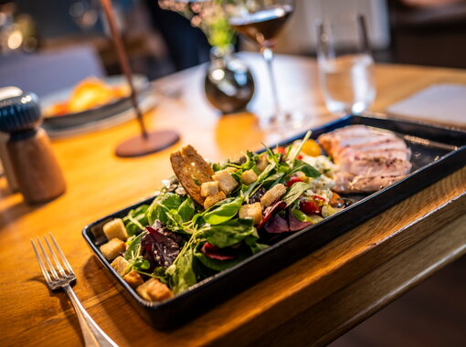 Nahaufnahme eines angerichteten Gerichts mit geschnittenem, gewürztem Steak, gemischtem Salat mit Cherrytomaten, Croutons und Dressing auf einem schwarzen Teller. Im Vordergrund ein Glas Rotwein auf einem Holztisch.