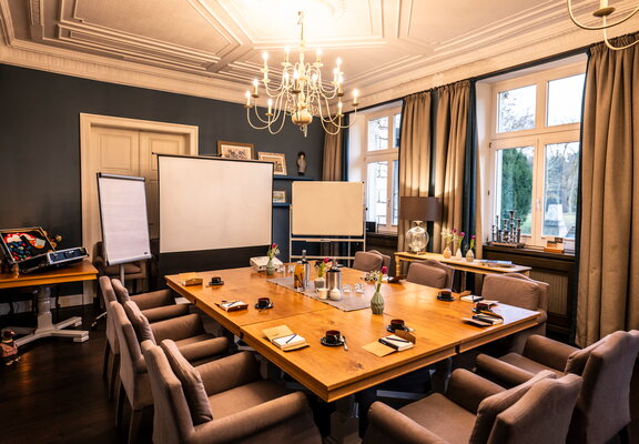 Ein gut ausgestatteter Konferenzraum mit einem großen Holztisch, umgeben von gepolsterten Stühlen. Auf dem Tisch liegen Notizblöcke, Stifte, Tassen und Untertassen. Im Raum befinden sich zwei Whiteboards und ein Flipchart-Ständer. Ein Kronleuchter hängt